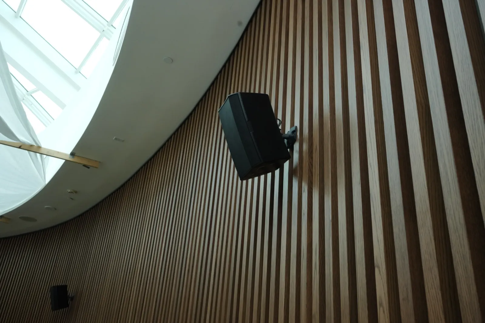 Black surface-mounted loudspeaker installed on a curved wood-slat wall beneath a skylight