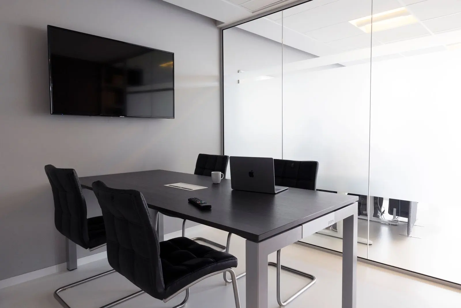 Small meeting room with a wall-mounted display, table and chairs behind a glass partition
