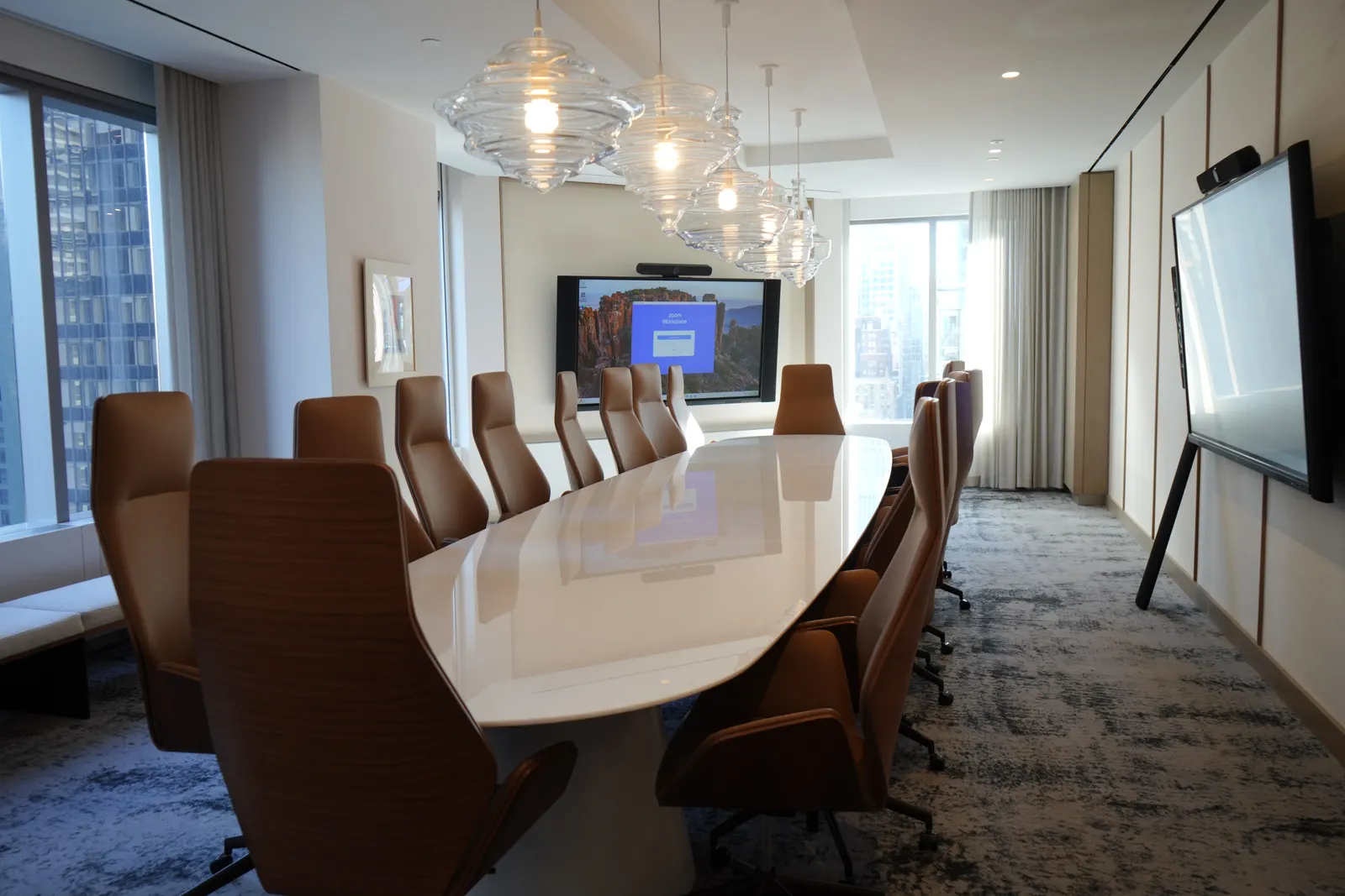 Boardroom with a long white table, leather chairs and dual wall displays on a video call, city skyline through the windows