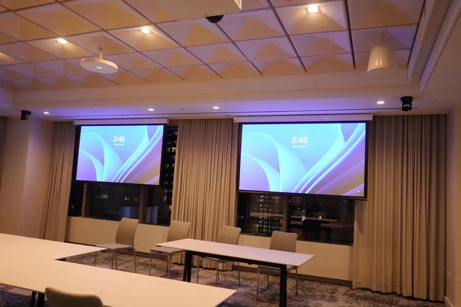 Training room with two projection screens above rows of tables, acoustic ceiling and city view at night